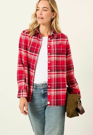 Red plaid flannel shirt with white and black accents, button-up front, long sleeves, and a chest pocket, paired with light blue jeans.
