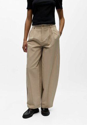 Femme portant un pantalon beige taille haute à jambes larges avec plis, un haut noir à manches courtes et des mocassins noirs, debout devant un fond blanc.