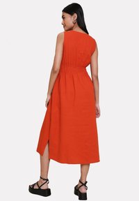 Vestido sin mangas de longitud media en una tela de color naranja brillante, con una cintura elástica y una abertura lateral. Combinado con sandalias negras de tiras.