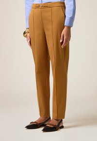 Pantalon beige avec une coupe ajustée, doté d'un pli avant, d'une ceinture avec passants et d'un petit détail de bouton rond. Porté avec des ballerines noires à bouts pointus.