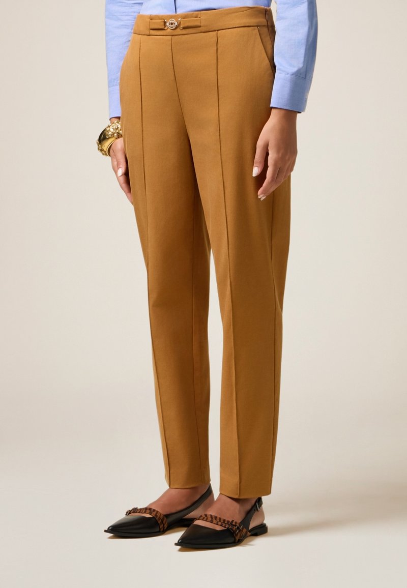 Pantalon beige avec une coupe ajustée, doté d'un pli avant, d'une ceinture avec passants et d'un petit détail de bouton rond. Porté avec des ballerines noires à bouts pointus.