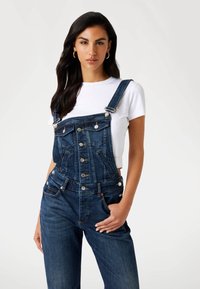 Dunkelblaue Denim-Latzhose mit strukturiertem Design, metallischen Knopf-Akzenten und Vordertaschen, kombiniert mit einem schlichten weißen T-Shirt.