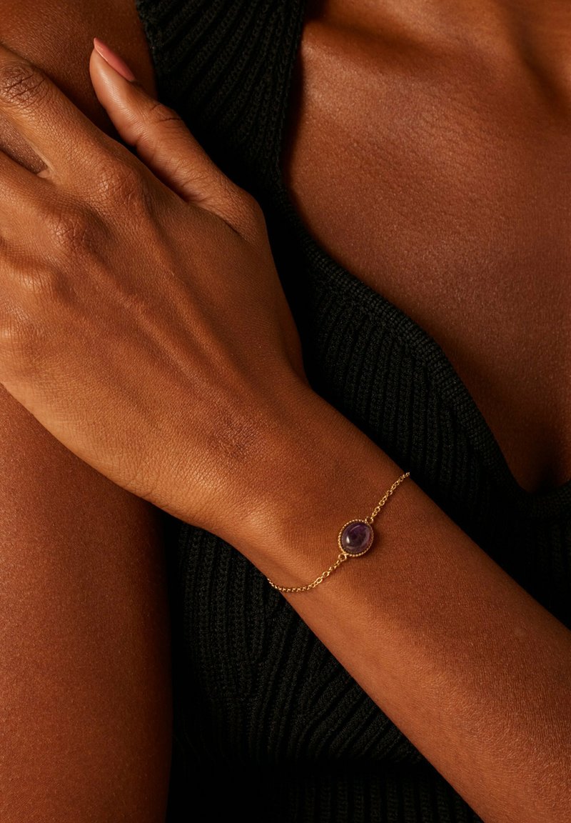 Gold chain bracelet featuring an oval amethyst stone, set in a textured bezel, worn on a brown skin tone with a black ribbed top.