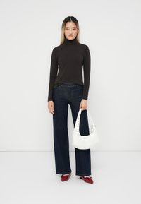 Brun polotröja, mörk denim utsvängda jeans, vit handväska med utskärningsdetalj, röda klackskor, enfärgad vit bakgrund.