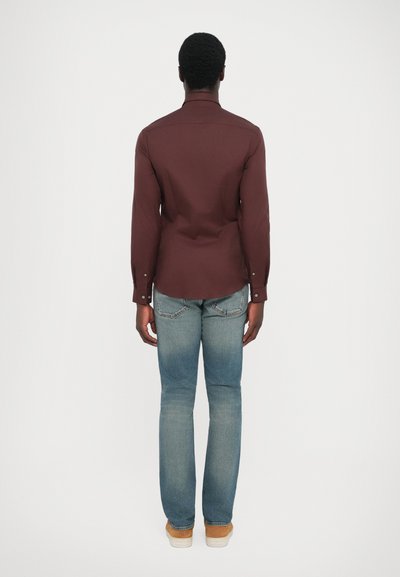 Camisa marrón de manga larga con corte recto, combinada con jeans de color azul claro. La camisa presenta un cuello estándar y puños abotonados.
