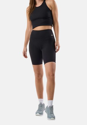 Kobieta w czarnym, bezrękawnym crop topie oraz czarnych, rowerowych szortach z wysokim stanem, białych skarpetkach i szarych sportowych butach na białym tle.