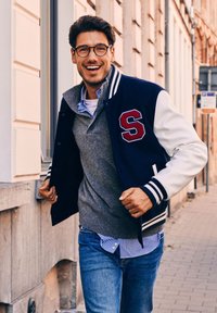 Collegejack in marineblauw met witte leren mouwen en de rode letter 'S.' Grijze gebreide sweater over een blauw geruit shirt. Draagt een blauwe spijkerbroek.