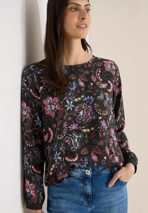 Vrouw draagt een donker bloesje met lange mouwen, gestoken in een blauwe spijkerbroek, staat met één hand in de zak en kijkt opzij.