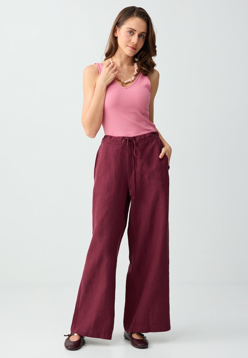 Femme portant un haut rose sans manches, un pantalon large bordeaux avec cordon, des ballerines foncées et un collier en coquillage, debout devant un fond clair.