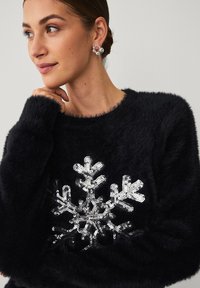 Zwarte trui van namaakbont met een zilveren pailletten sneeuwvlokontwerp op de voorkant, voorzien van een ronde hals en lange mouwen.