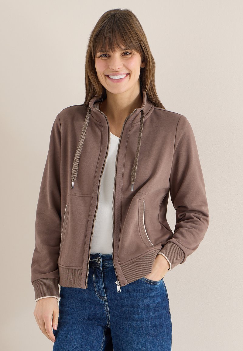Femme souriante portant une veste marron à fermeture éclair avec cordons et un haut blanc, ainsi qu'un jean bleu, posant devant un fond beige uni.