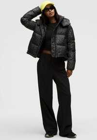 Jachetă de puf cu imprimeu leopard negru, design scurt, asortată cu un top negru și pantaloni negri largi. Accesoriile includ o șapcă galbenă strălucitoare.