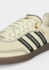 Κρεμ αθλητικό παπούτσι Adidas Samba με μαύρα ραβδωτά ράμματα, λευκά κορδόνια, χρυσά κουμπώματα και λαστιχένια σόλα, που δείχνει πλευρικές και επάνω λεπτομέρειες.