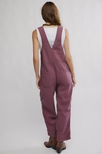 Bordeauxrote Baumwoll-Overalls mit weitem Bein, V-förmigem Rückendesign und zwei vorderen Taschen. Getragen über einem weißen Tanktop. Braune Stiefeletten vervollständigen den Look.