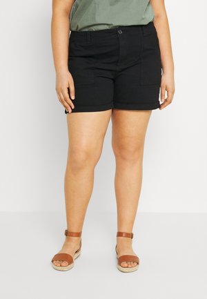 Personne portant un short noir, un haut vert et des sandales marron à bride à la cheville, debout sur un fond blanc uni.