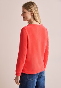 Koraalkleurig langslavend shirt met een gemêleerd uiterlijk, ronde halslijn en ribgebreide manchetten. Gecombineerd met blauwe spijkerbroek.