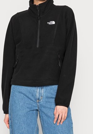 Persoon gekleed in een zwarte fleecejas van The North Face met halve ritssluiting en lichtblauwe spijkerbroek, staand tegen een effen achtergrond.