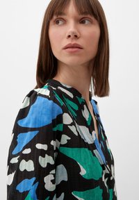 Blouse noire avec des motifs abstraits colorés en bleu, vert et blanc. Présente un décolleté en V et un tissu doux et texturé. Manches longues.