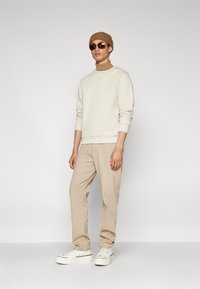 Felpa beige con polsini a costine, abbinata a pantaloni beige chiaro. Indossata con un berretto di maglia marrone e occhiali da sole scuri, completata da sneakers bianche.