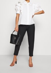 Žena v bílé volánkové blůze s krátkým rukávem, černých pruhovaných kalhotách, světlých sandálech na podpatku, držící černou kabelku se stříbrnou sponou.