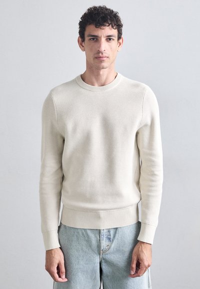 Maglione lavorato a maglia crema con colletto rotondo, polsini e orlo a coste, abbinato a jeans blu chiaro. Presenta un tessuto testurizzato.