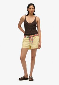 Top in maglia marrone, pantaloni shorts gialli con una cintura a fantasia e sandali scivolati marroni. Tessuti lisci, vestibilità casual.