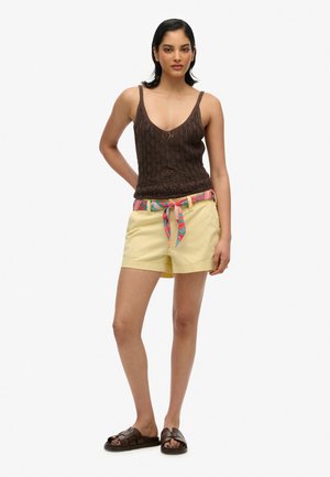 Top in maglia marrone, pantaloni shorts gialli con una cintura a fantasia e sandali scivolati marroni. Tessuti lisci, vestibilità casual.