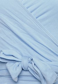 Ljusblå omslagstopp i mjukt tyg, med diagonal ribbad struktur och ett knytband vid midjan för säker stängning.