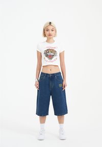 White cropped t-shirt featuring a colorful tiger graphic, paired with loose, knee-length blue denim shorts with embroidery detail. White sneakers.
