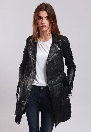 Black leather jacket featuring a long design, diagonal zipper closure, quilted shoulder accents, and bold stitching. Paired with a white shirt and blue jeans.