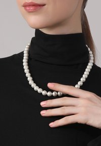 Perlenkette mit runden, glänzenden weißen Perlen, getragen über einem schwarzen Hochkragenoberteil. Hand ruht sanft auf der Kette.