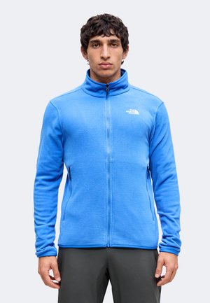 Άνδρας που φοράει ένα φωτεινό μπλε μπουφάν fleece με φερμουάρ, ψηλό γιακά και τσέπες με φερμουάρ στα πλάγια, κοιτάζει μπροστά.