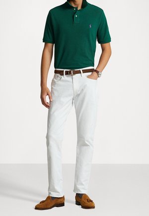 Mann trägt ein dunkelgrünes Polohemd, weiße Jeans mit brauem Gürtel, braune Quastenleder Schuhe und eine Armbanduhr, posierend mit einer Hand in der Tasche.