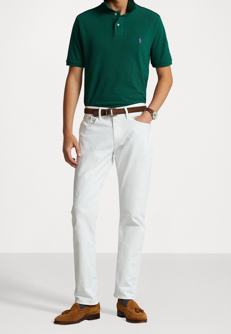 Homme portant un polo vert foncé, un jean blanc avec une ceinture marron, des mocassins à franges bruns et une montre au poignet, posant avec une main dans sa poche.