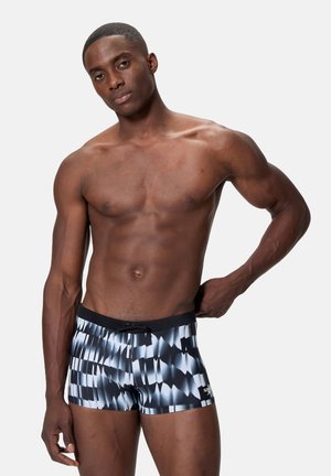 Muscular man wearing black and white patterned swim shorts stands against a plain light background, with one hand resting on his hip.