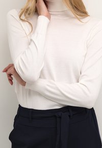 Pull à col roulé blanc en maille côtelée, fabriqué en tissu tricoté doux, avec des manches longues et des poignets ajustés, associé à un pantalon taille haute bleu marine.