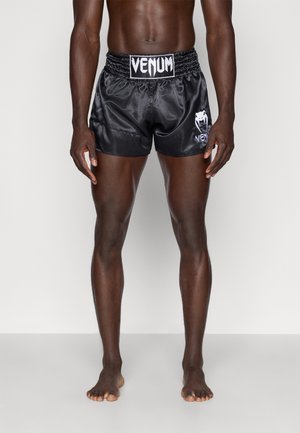 Mandlig model iført sorte satin Venum fight shorts, der står barfodet mod en ensfarvet hvid baggrund.