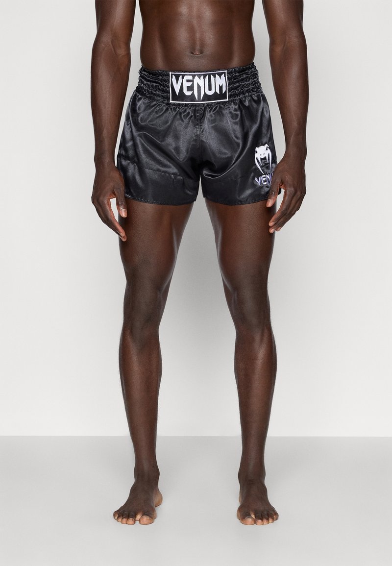 Modèle masculin portant un short de combat Venum en satin noir, debout pieds nus sur un fond blanc uni.