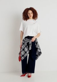 Νεαρή γυναίκα με σγουρά μαλλιά που φοράει λευκό μπλουζάκι, σκούρο τζιν με φαρδιά πόδια, πουκάμισο καρό δεμένο στη μέση, κόκκινα τακούνια και κρατά κόκκινη τσάντα.