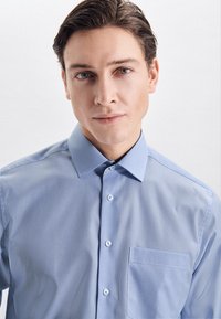 Camicia a righe blu chiaro con colletto a punta, tasca frontale e chiusura a bottoni. Tessuto liscio e vestibilità su misura.