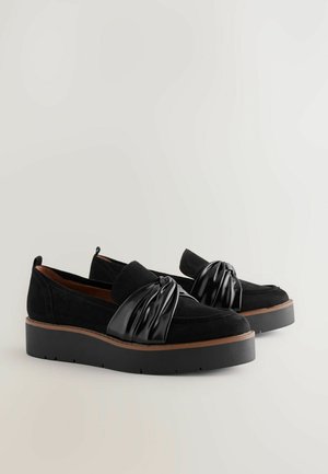 Schwarze Wildlederschuhe mit einem glänzenden schwarzen plissierten Akzent, Plateausohle und kontrastierendem braunem Rand; Slip-On-Design und runde Form.