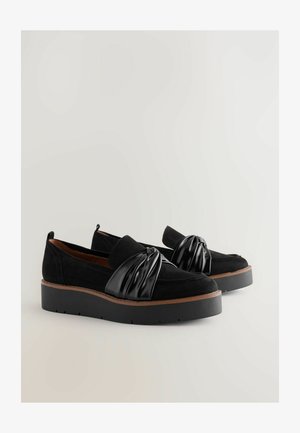 Black suede shoes with a shiny black pleated accent, platform sole, and contrasting brown edge; slip-on design and rounded shape.