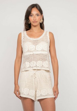 Femme portant un haut sans manches en crochet blanc et un short assorti avec des motifs circulaires, debout devant un fond clair uni.