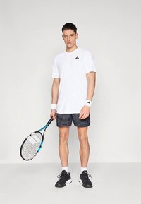 Λευκό πουκάμισο με κοντά μανίκια και μαύρο λογότυπο, μαύροι σορτς με σχέδιο, λευκές βραχιόνες, κρατώντας μπλε και λευκή ρακέτα τένις, μαύρα αθλητικά παπούτσια.