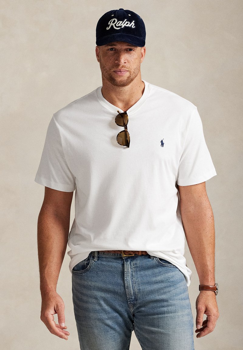 Bílé bavlněné tričko s malým modrým logem, kombinované s světlými modrými džíny. Doplňky zahrnují sluneční brýle a tmavě modrou čepici s bílým nápisem.