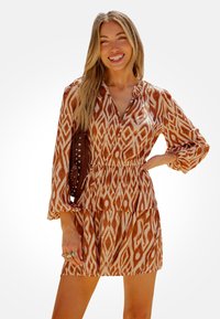 Abito marrone e crema con fantasia, maniche lunghe, scollatura a V e vita elastica. La modella tiene una borsa marrone con borchie.