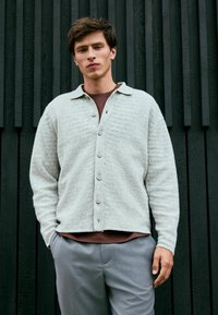 Hellgrauer Herringbone-Cardigan mit Kragen,Knopfverschluss und langen Ärmeln, über einem braunen Hemd getragen und mit grauen Hosen kombiniert.