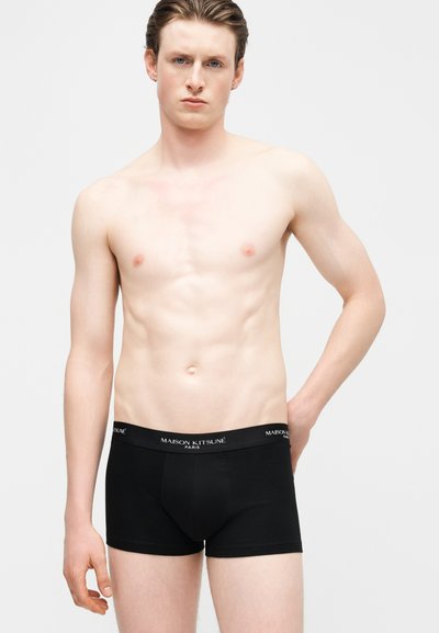 Νέος άνδρας με ανοιχτόχρωμο δέρμα και ανοιχτά μαλλιά που φοράει μαύρα boxer Maison Kitsuné, στέκεται μπροστά σε απλό ανοιχτό φόντο με το ένα χέρι πίσω από την πλάτη.