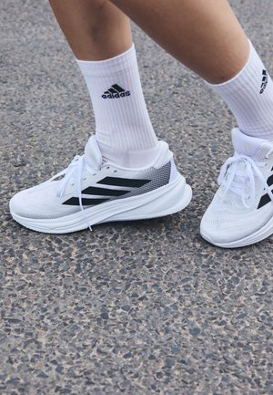 Osoba stojící na asfaltovém povrchu, obutá v bílých běžeckých botách Adidas a odpovídajících bílých ponožkách Adidas.