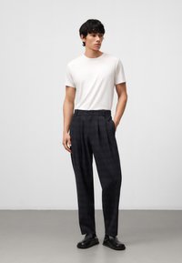 Witte t-shirt met korte mouwen, gecombineerd met donker geruite hoge taille broek. Het model staat op een grijze vloer, met de handen in de zakken, en draagt zwarte schoenen.